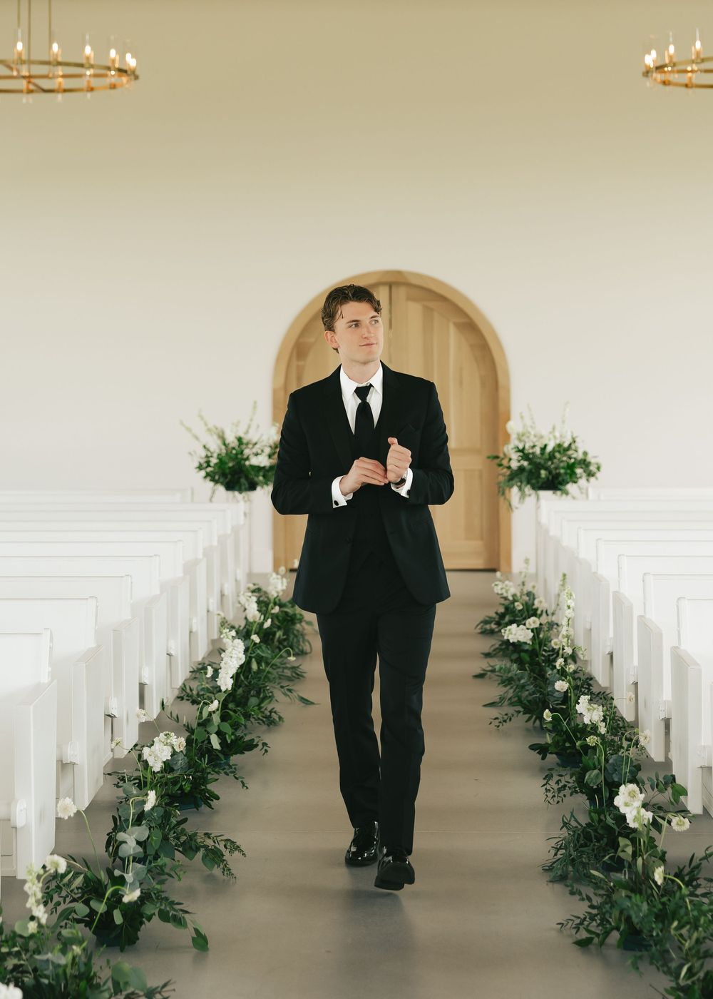 Dapper groom in a black suit walking down a white chapel aisle lined with lush greenery floral arrangements.