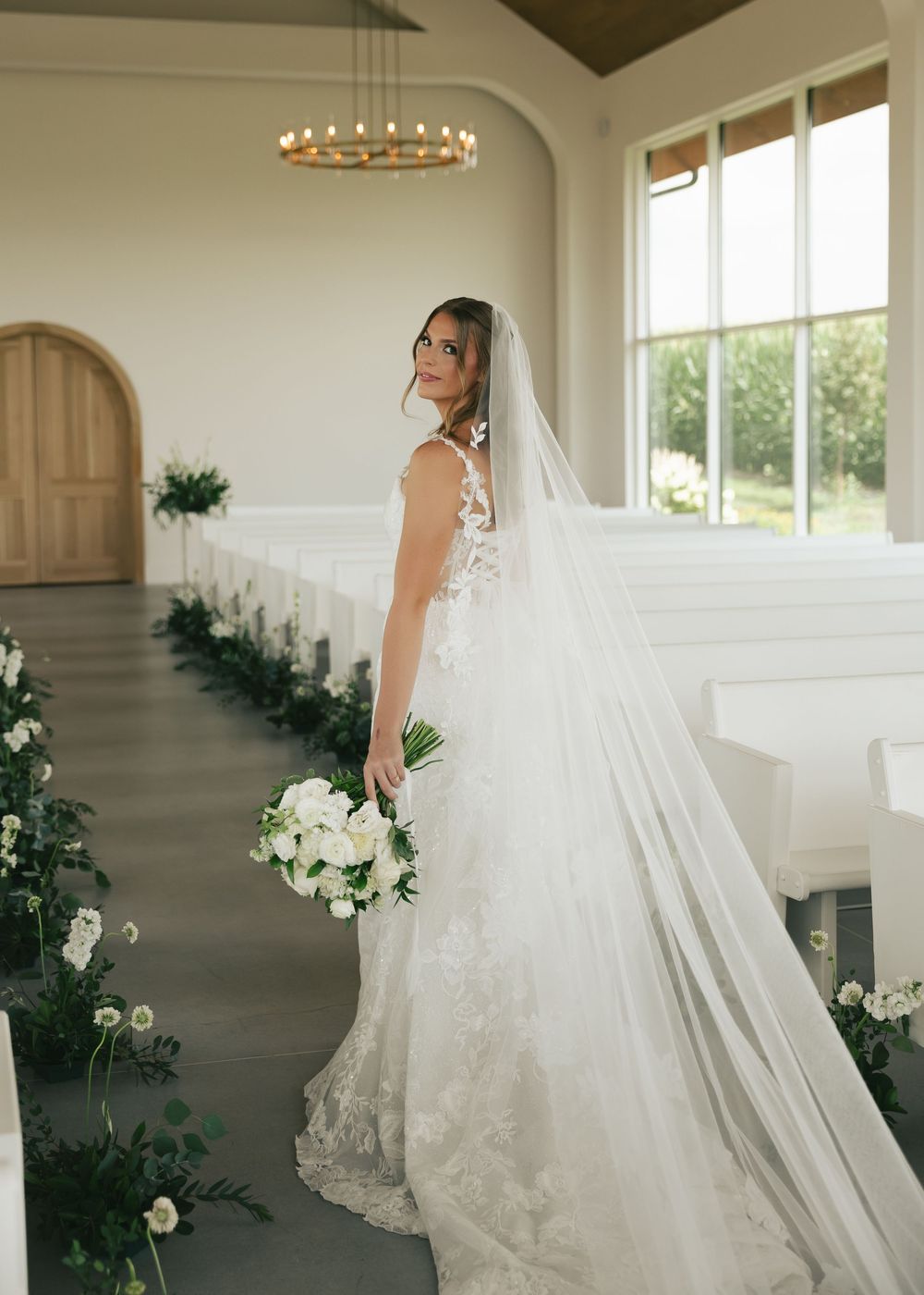 Bride in lace wedding gown and long veil holds white bouquet in elegant white chapel aisle with greenery decor.