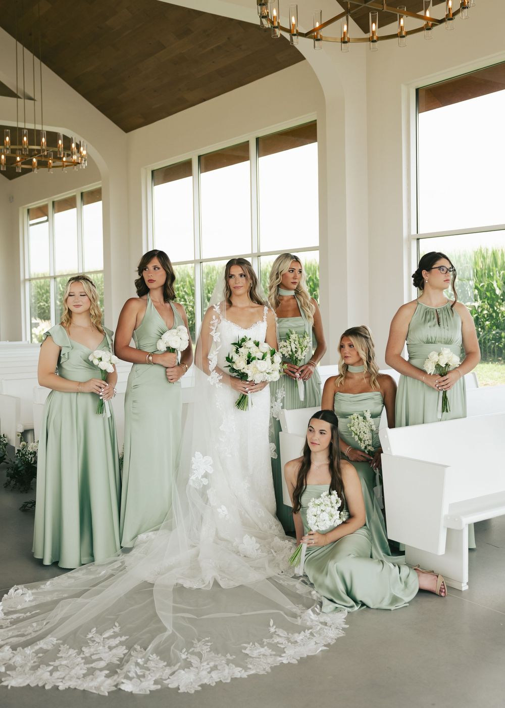 Bride in white lace gown with six bridesmaids in sage green dresses holding white bouquets in elegant chapel.