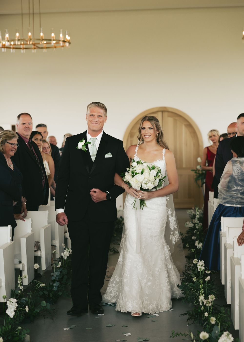 Bride in lace gown walks down aisle with father at elegant white wedding ceremony with floral arrangements.