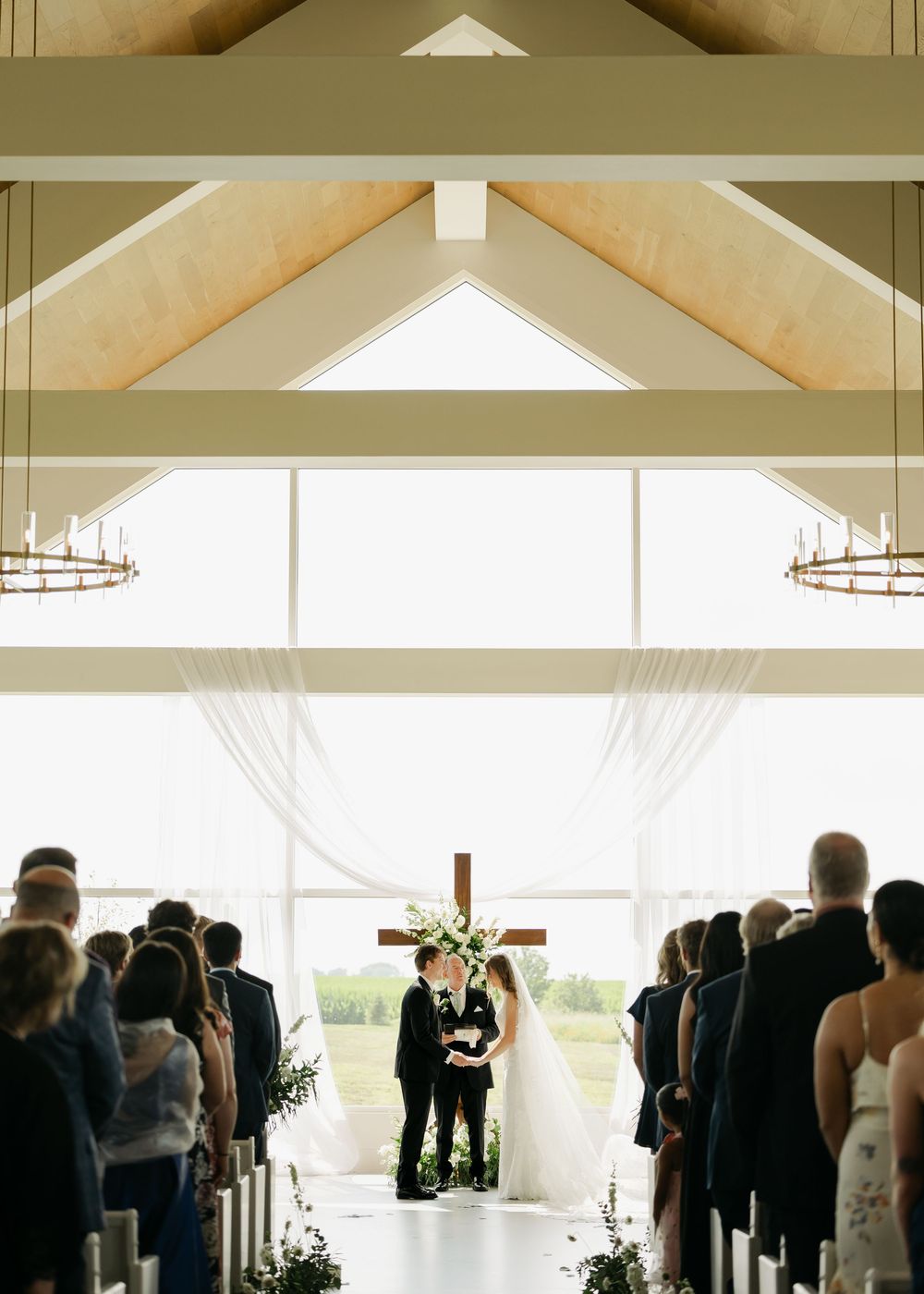 Couple exchanges vows at elegant barn wedding ceremony with wooden cross, sheer draping, and guests seated in aisle rows.