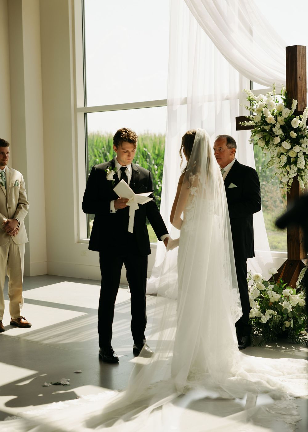 Groom reads heartfelt vows to bride in a sunlit chapel with white floral arrangements and sheer draping.