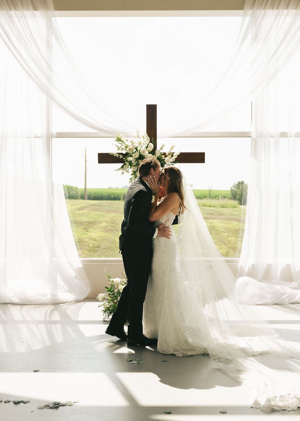 Bride and groom share first kiss at altar with wooden cross, white drapes, and scenic window backdrop.