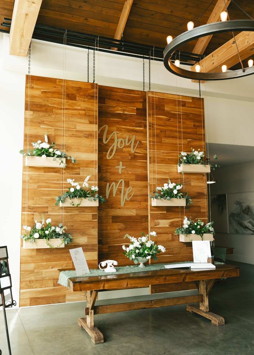 Rustic wooden wall wedding display with hanging floral boxes and a "You & Me" acrylic sign above a farmhouse table.