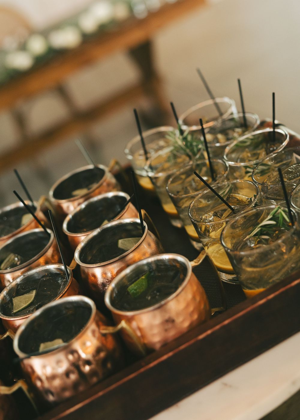 Copper Moscow mule mugs and cocktail glasses with black straws arranged on a wooden tray at a wedding reception.