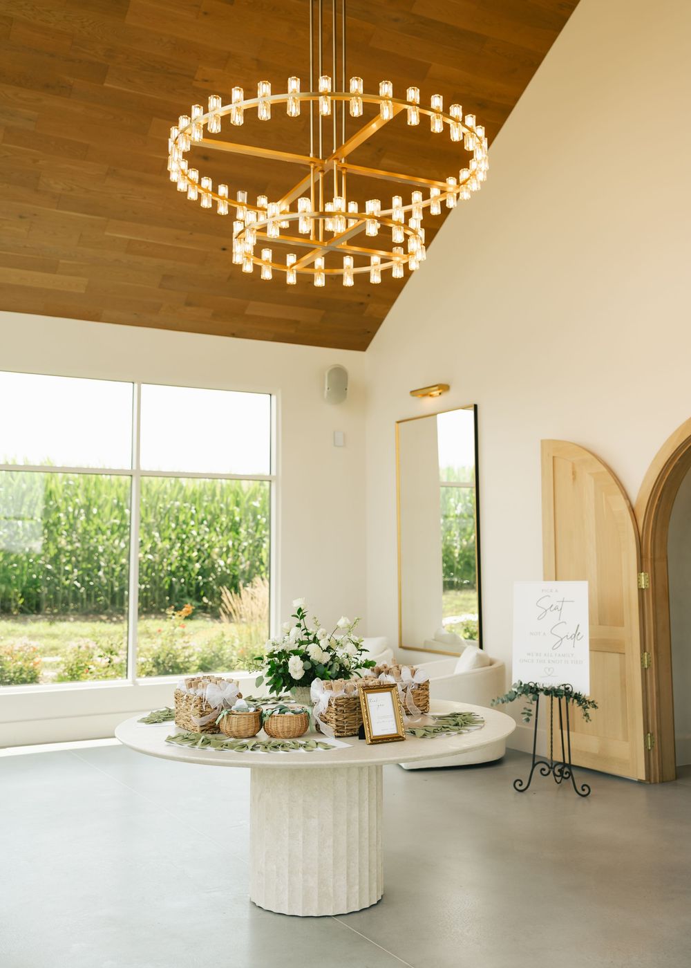 Elegant wedding venue interior with tiered gold chandelier, white pedestal table with floral arrangements, and floor-to-ceiling windows.