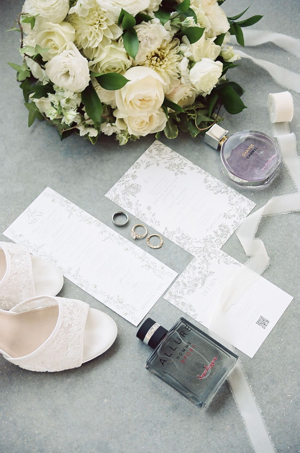 Elegant wedding flat lay with white roses bouquet, rings, Chanel perfume, bridal heels, and floral invitations.