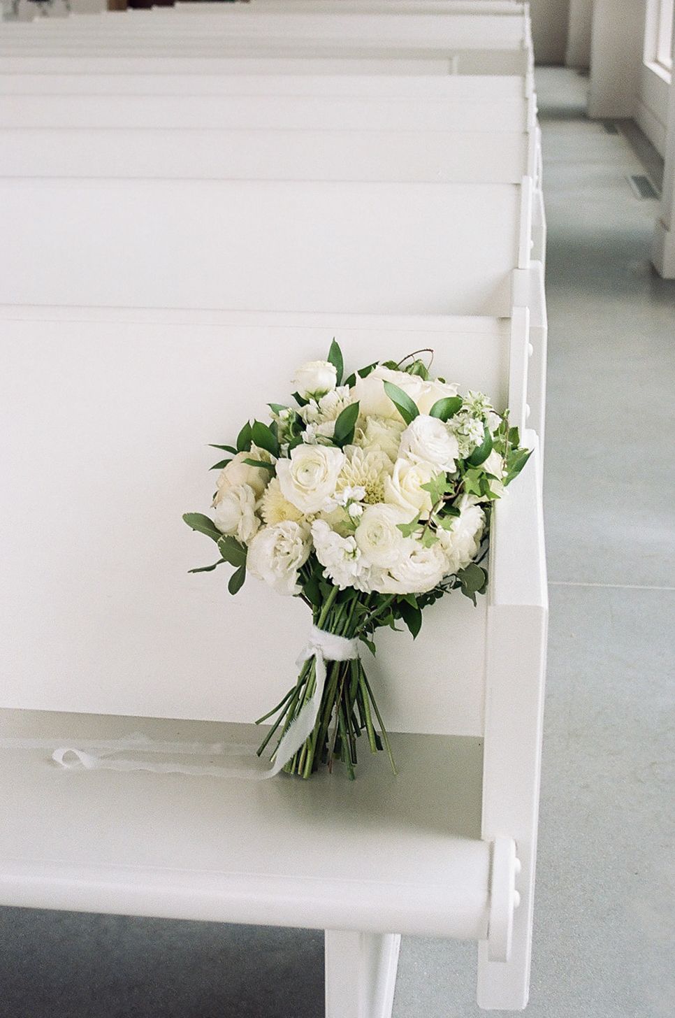 White wedding bouquet with roses and greenery resting on a white church pew.