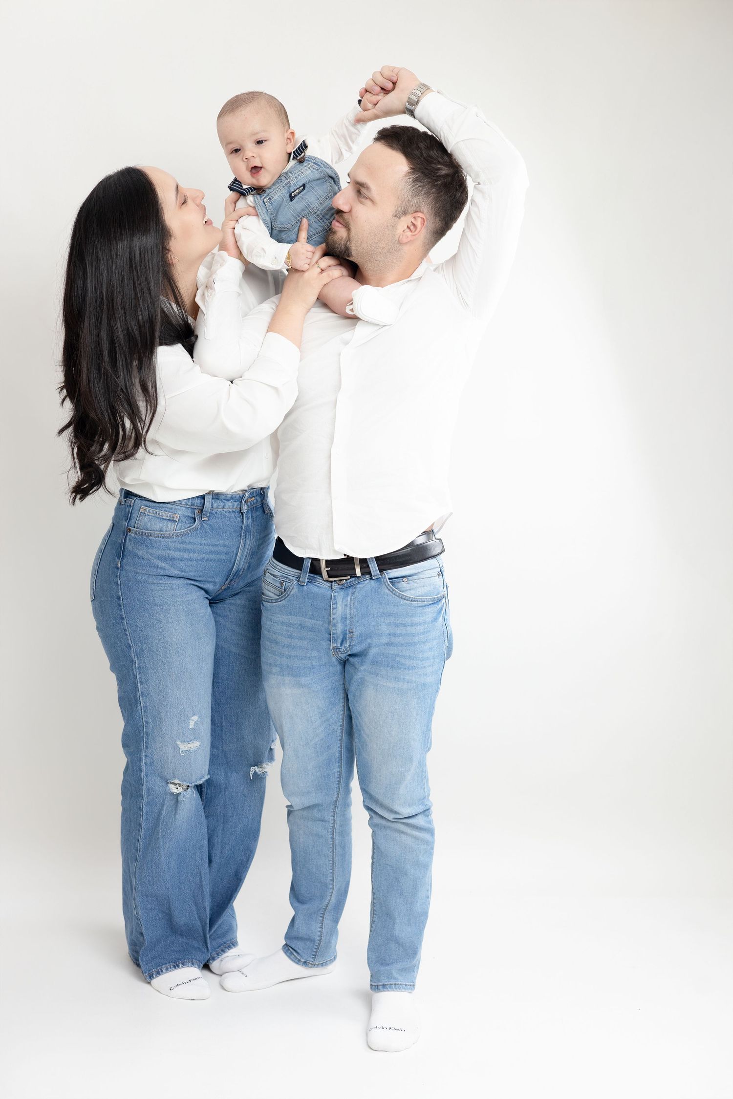 A family in matching white tops and blue jeans poses playfully against a white backdrop in a series of candid shots.