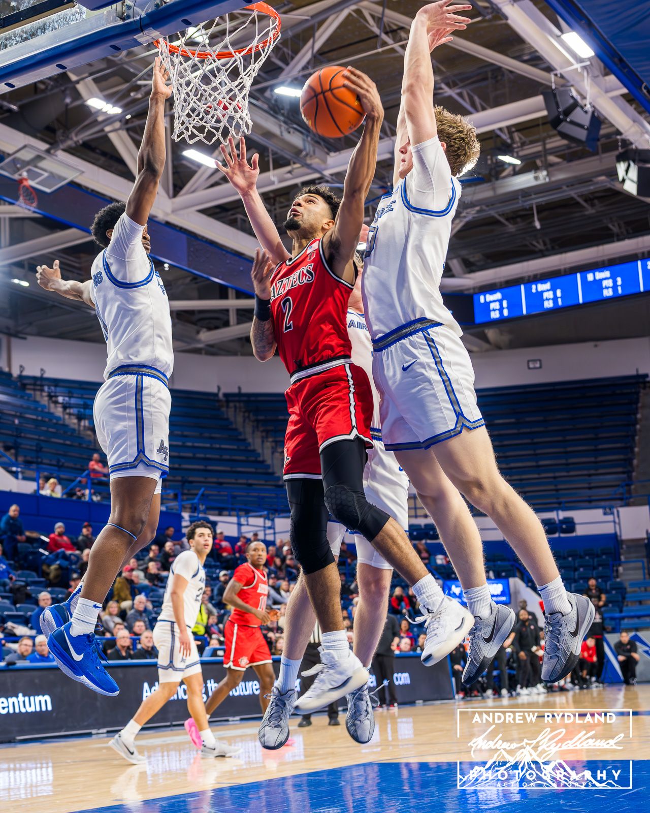 Basketball players leap for the ball during an intense game in a blue-lit indoor arena.