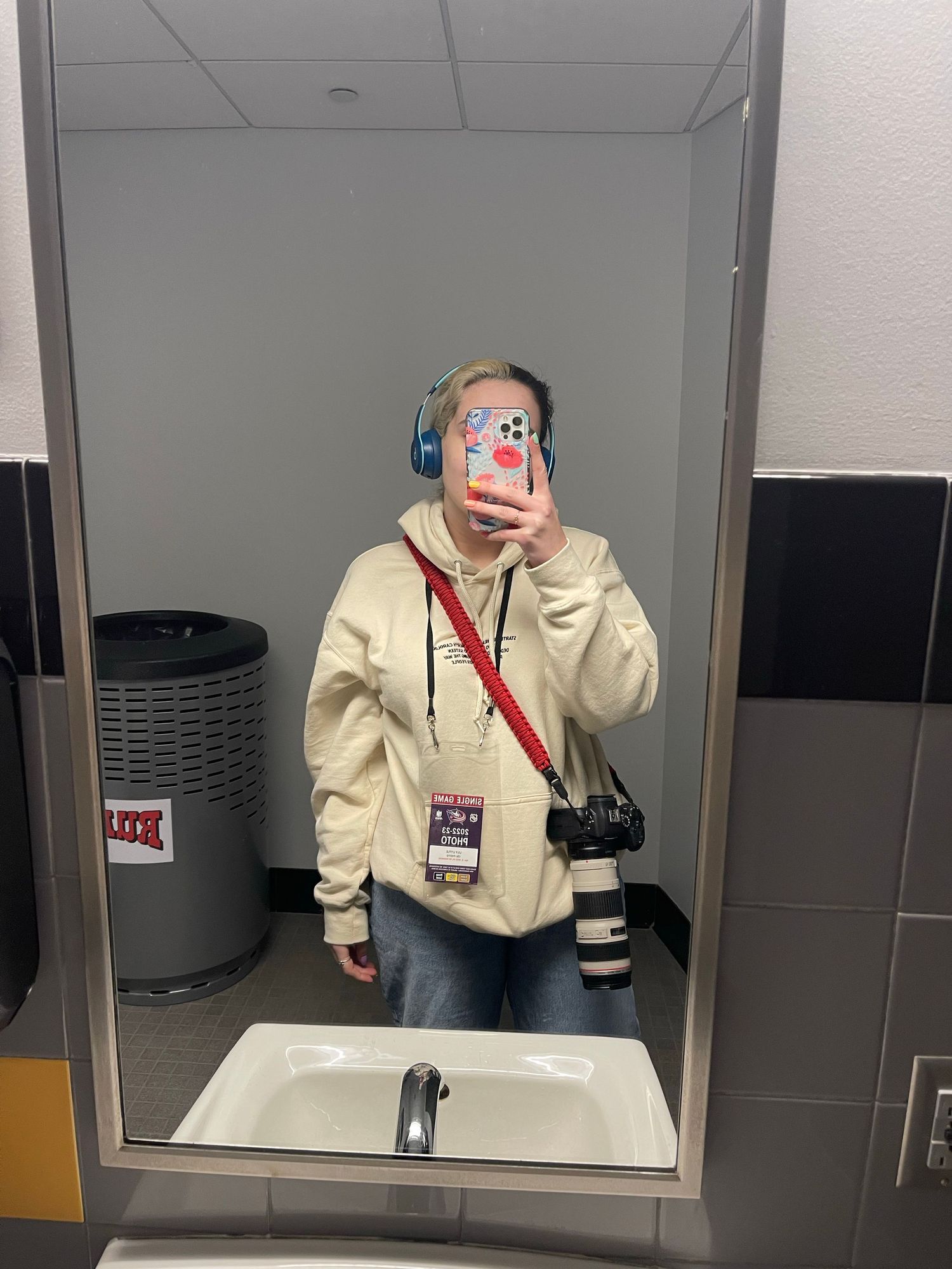 A photographer in a cream hoodie takes a mirror selfie in a bathroom while wearing headphones and carrying camera gear.