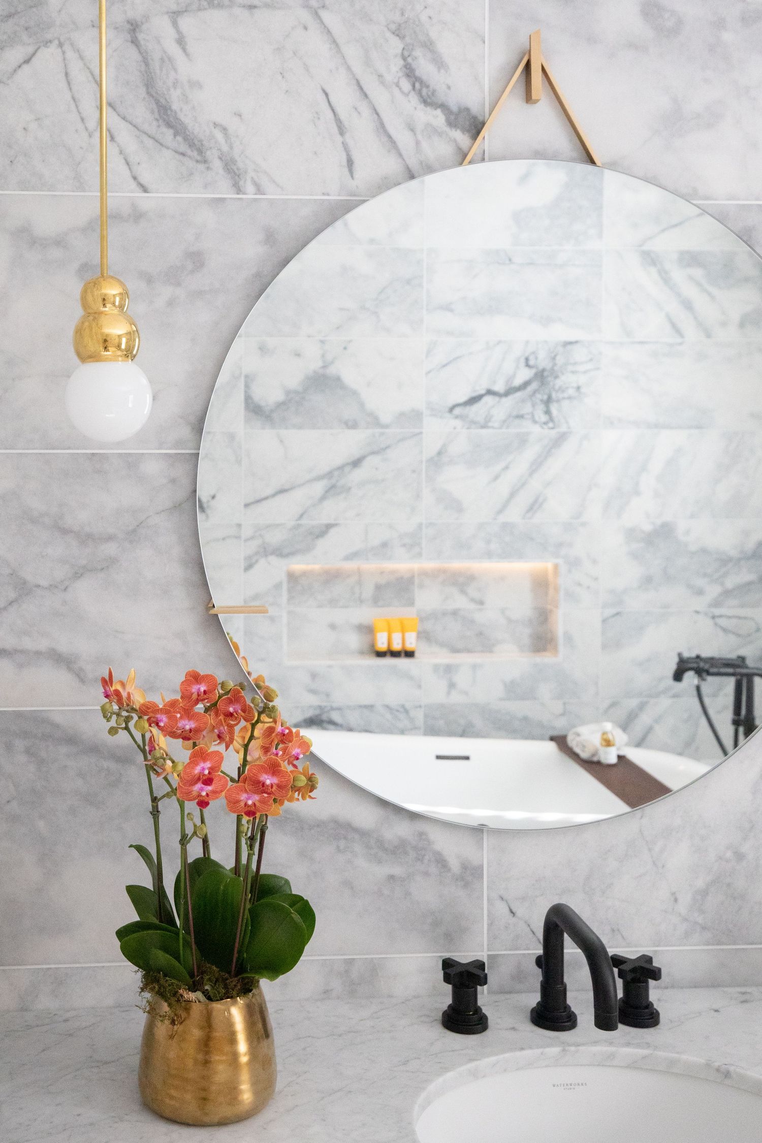 Modern marble bathroom with oval mirrors, gold accents, and pink orchids in a metallic planter against white walls.