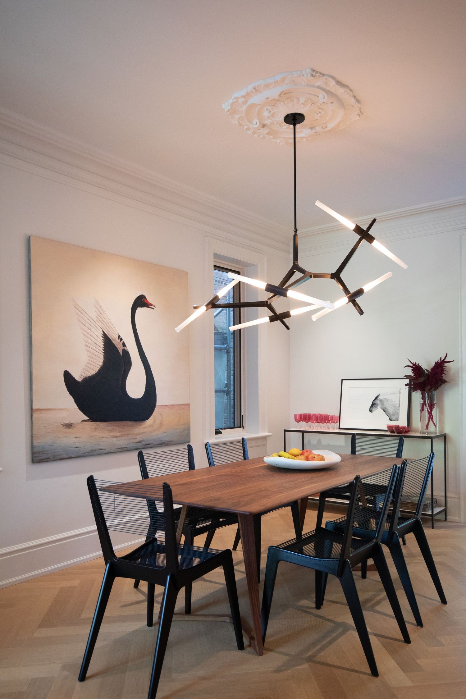 Modern dining room with wooden table, black chairs, swan artwork, and geometric pendant light fixture against white walls.