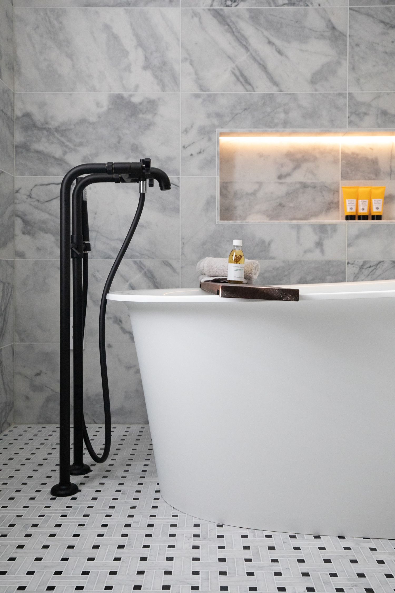 Modern freestanding bathtub with matte black floor-mounted tub filler against marble walls and geometric tile flooring.
