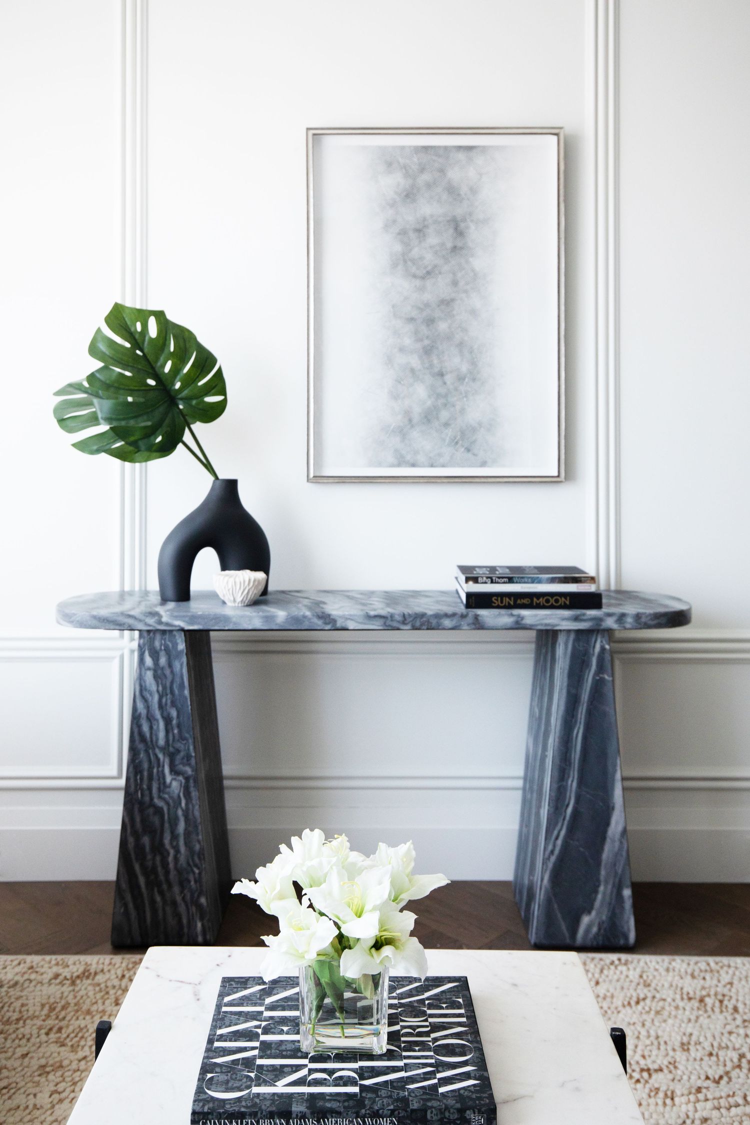 Modern console table vignette with marble top, black base, and monstera leaf in vase against white paneled wall.