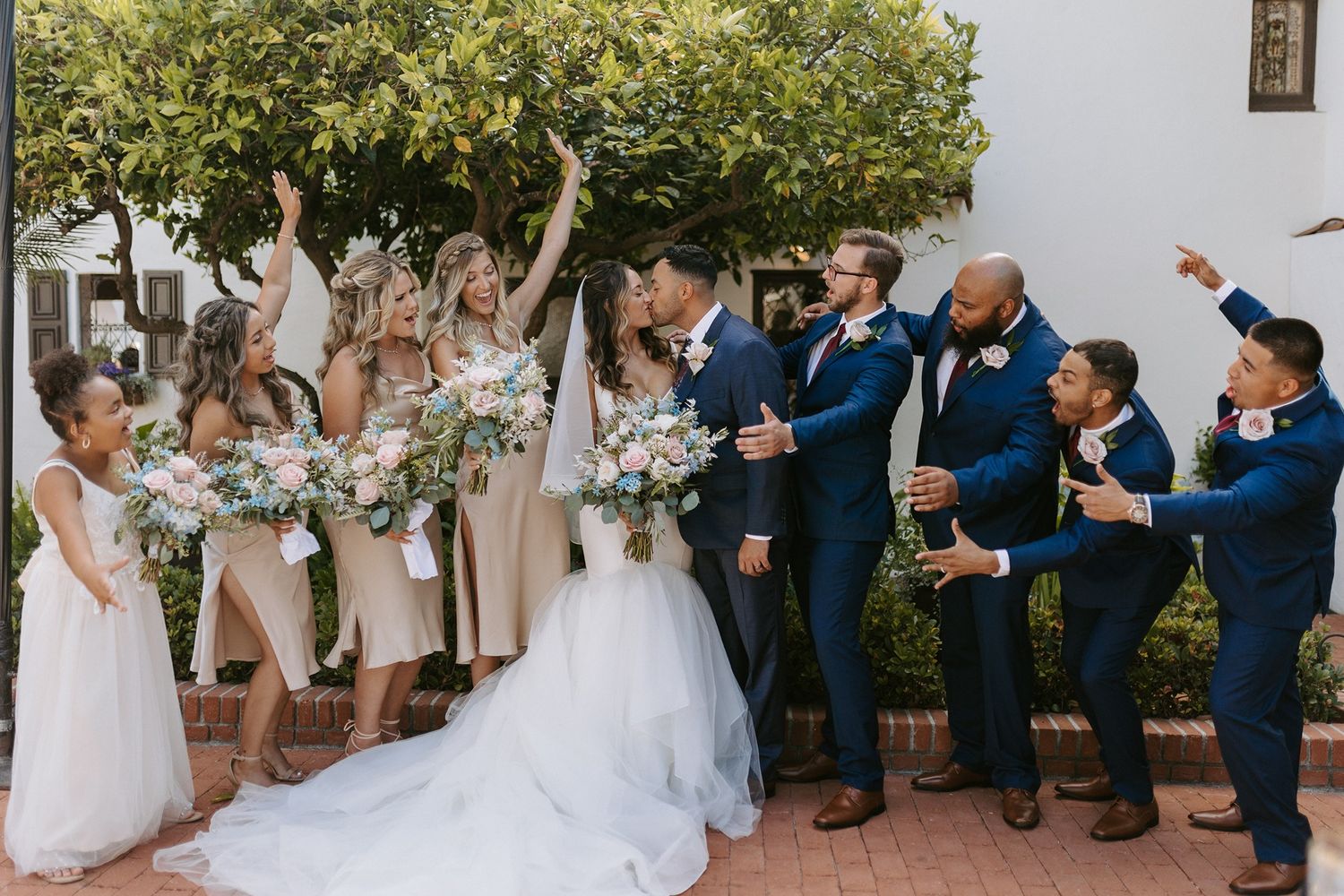 Wedding party celebrates as bride and groom kiss, bridesmaids in champagne gowns and groomsmen in navy suits cheering outdoors.