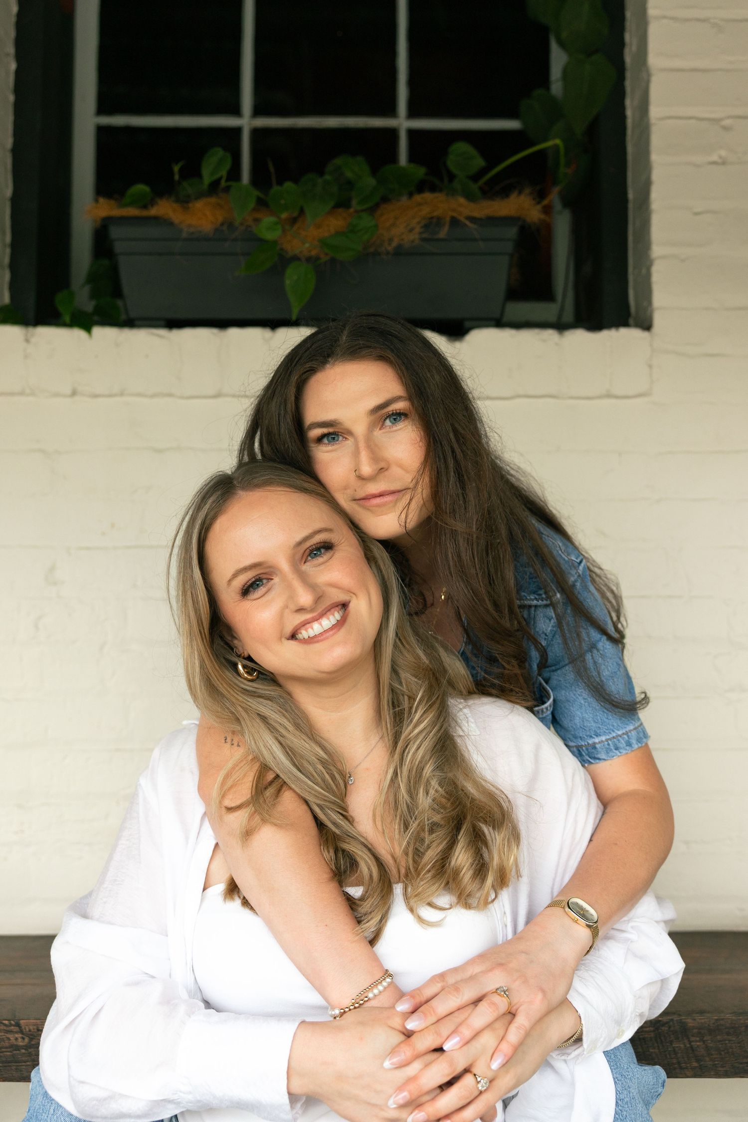 Two smiling women pose together on a porch, one hugging the other from behind, against a white brick wall.
