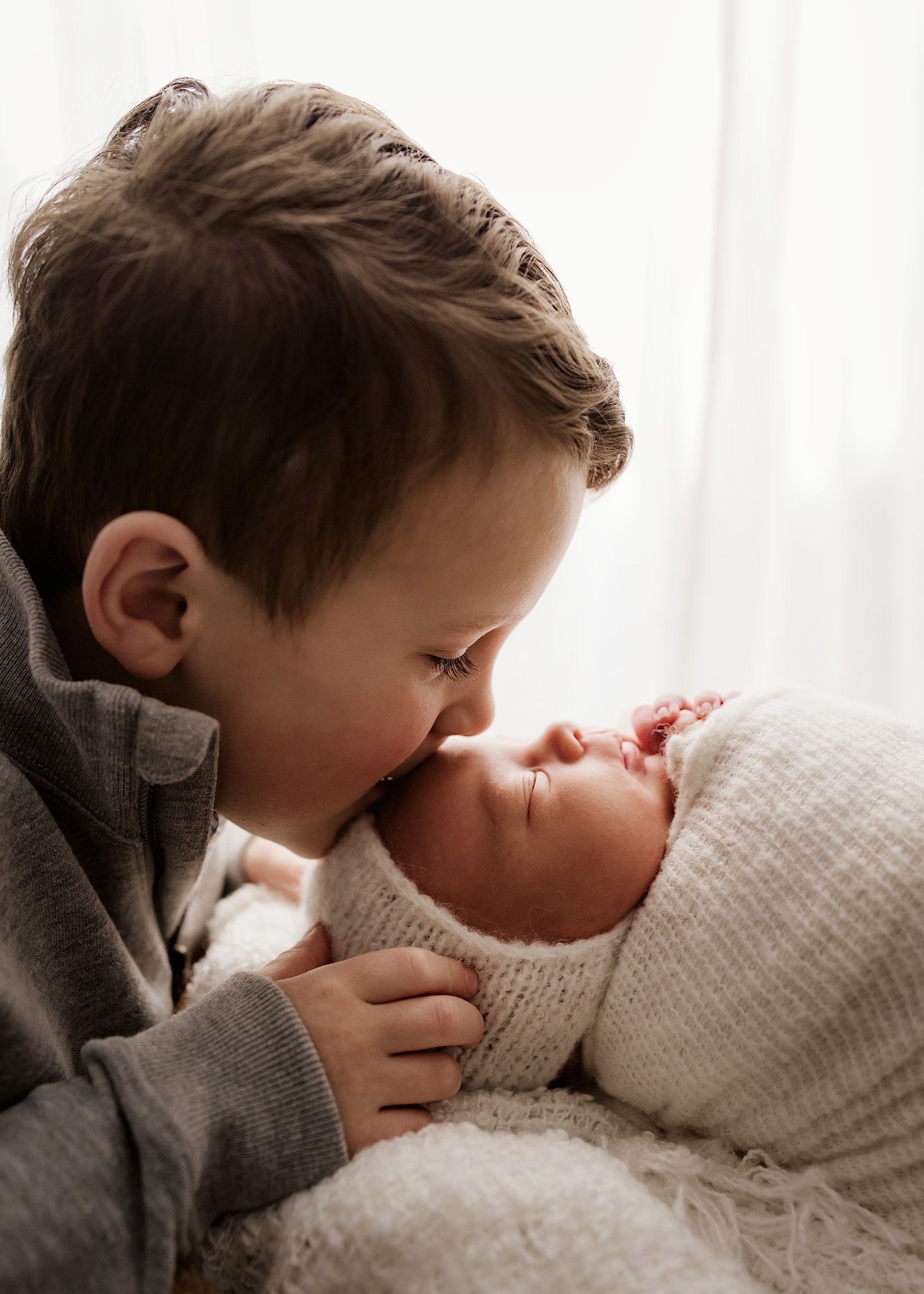 Young boy gently kissing sleeping newborn sibling wrapped in cream knit blanket.