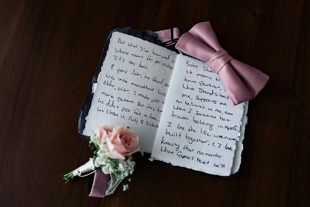 Open wedding vow book with handwritten notes beside a pink bow tie and boutonniere on a dark wood surface.