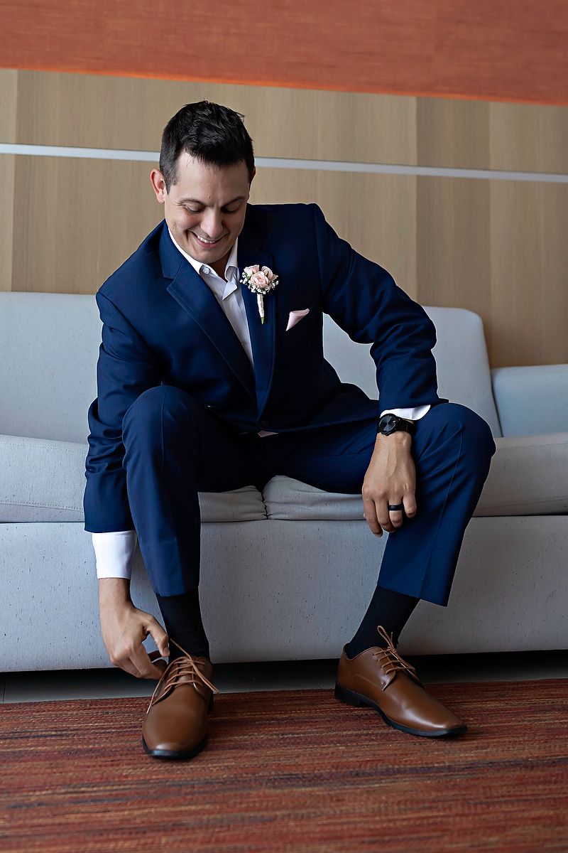 Smiling groom in navy blue suit with boutonniere tying brown dress shoes while seated on gray sofa.