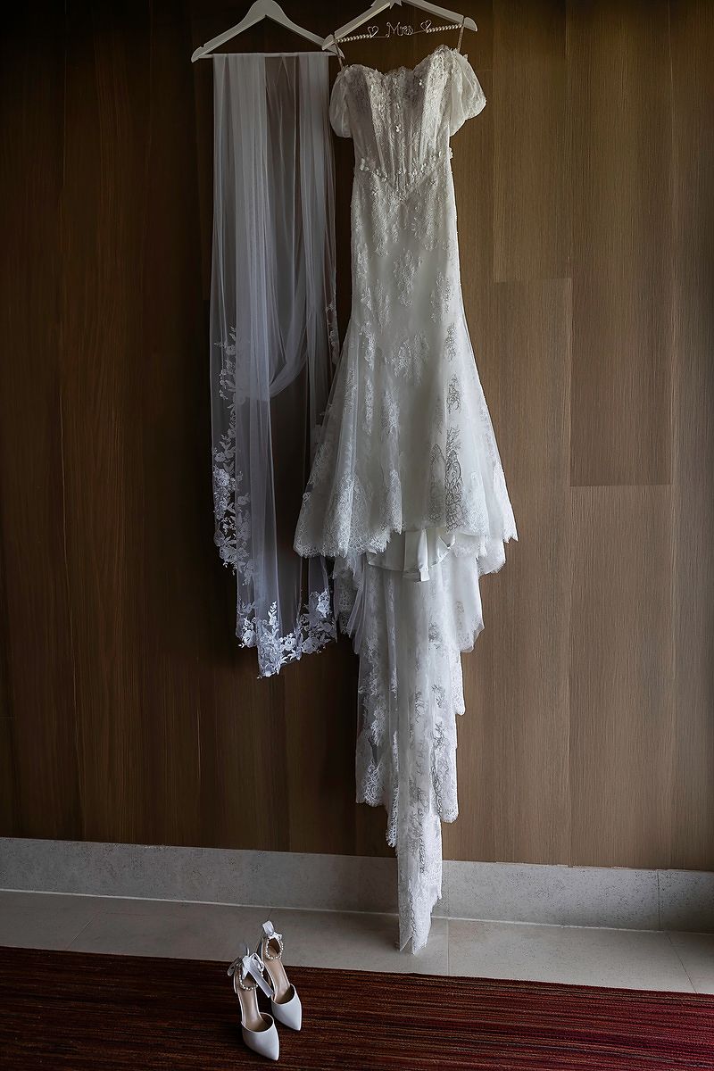 Elegant lace wedding dress and veil hanging on wooden hangers with white bridal heels below.