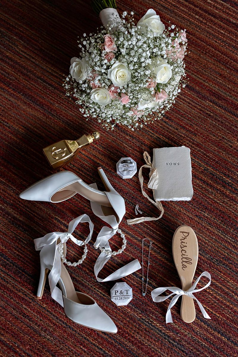 Flat lay of bridal accessories including white heels, bouquet, perfume, and personalized items on red carpet.