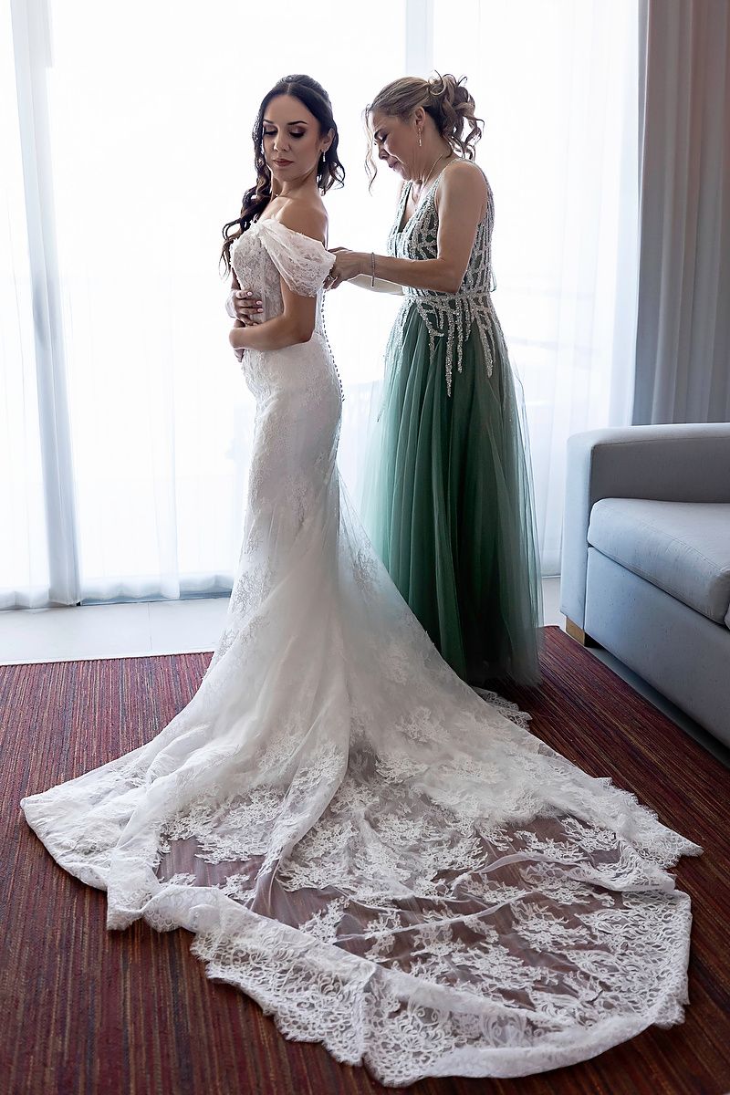 Bride in elegant lace mermaid wedding gown being assisted by bridesmaid in green dress near window.
