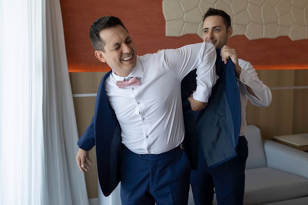 Two groomsmen in navy blue suits laugh while helping each other get dressed before a wedding ceremony.