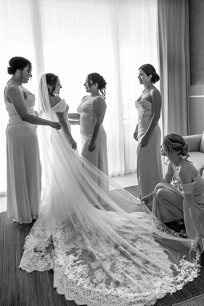 Black and white photo of bride with bridesmaids adjusting her lace cathedral veil before the ceremony.