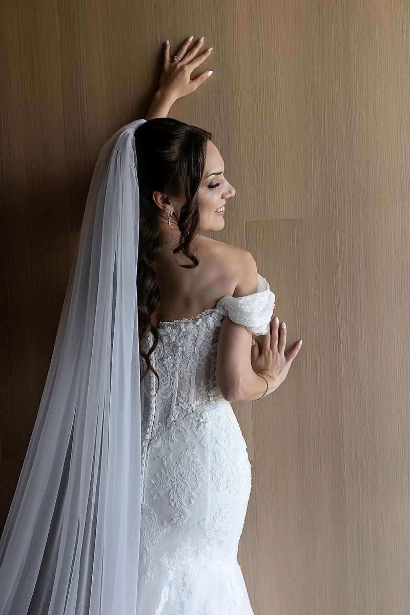 Elegant bride in off-shoulder lace mermaid gown with long veil poses gracefully against a wooden wall.