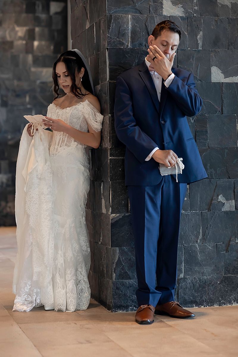 Bride in lace gown and groom in navy suit share private vows exchange moment, standing back-to-back against stone wall.
