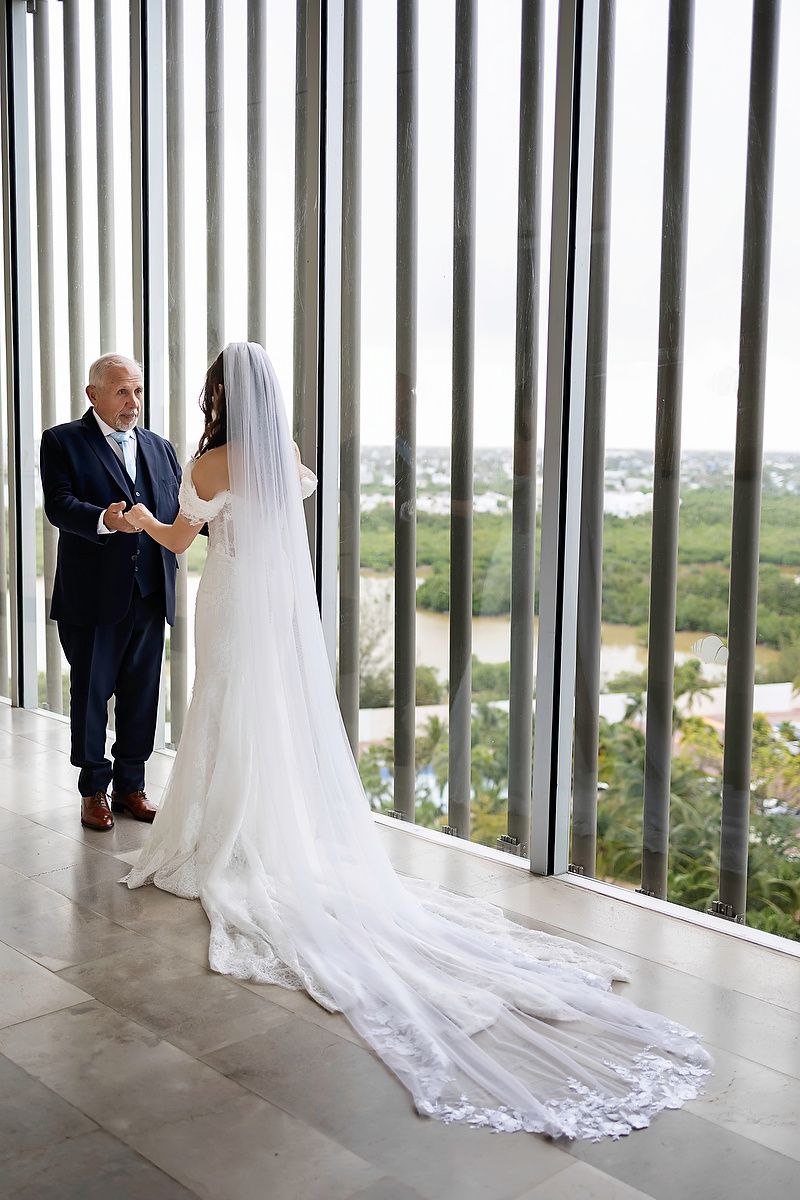Bride in white gown with long veil holds hands with man in navy suit near floor-to-ceiling windows overlooking lush scenery at Dreams Vista.