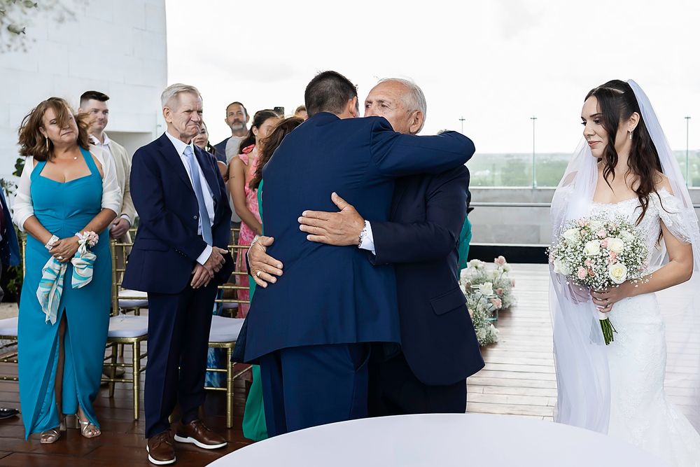 Groom hugging father-in-law at outdoor wedding ceremony while bride in white gown holds bouquet nearby.