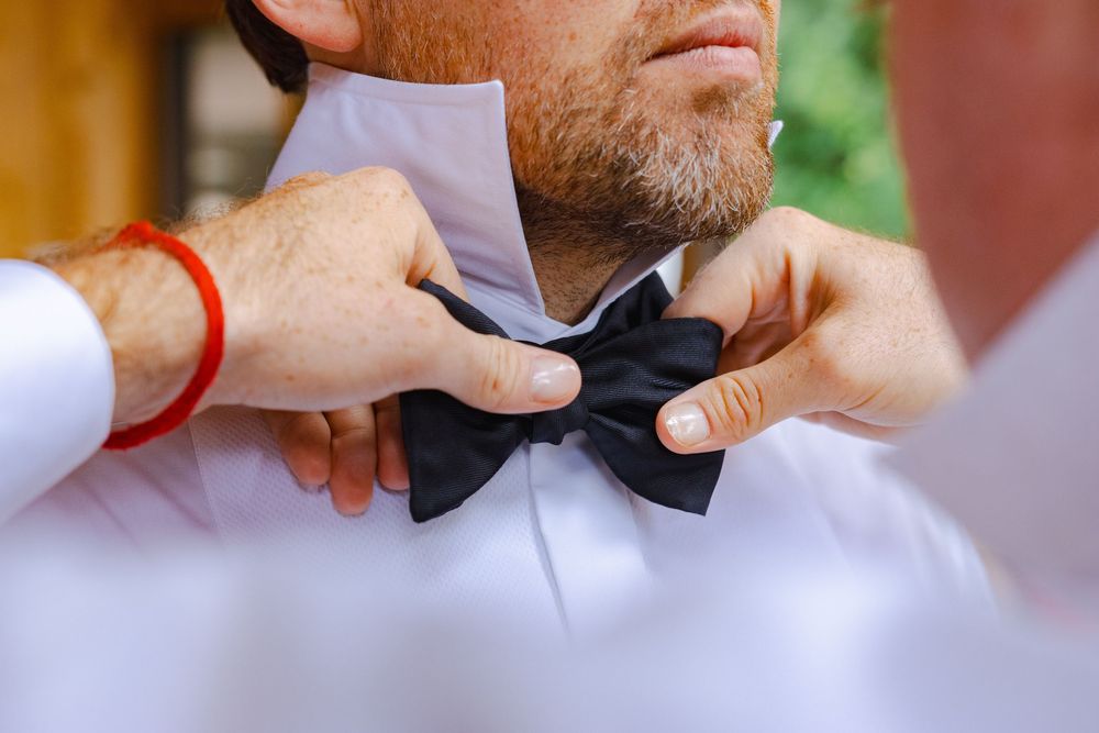 A man with a beard is adjusting his bowtie in preparation for a formal event.