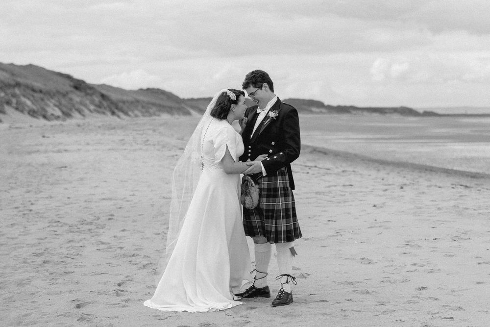 A couple in wedding attire shares an intimate moment on a scenic beach with mountains in the background.