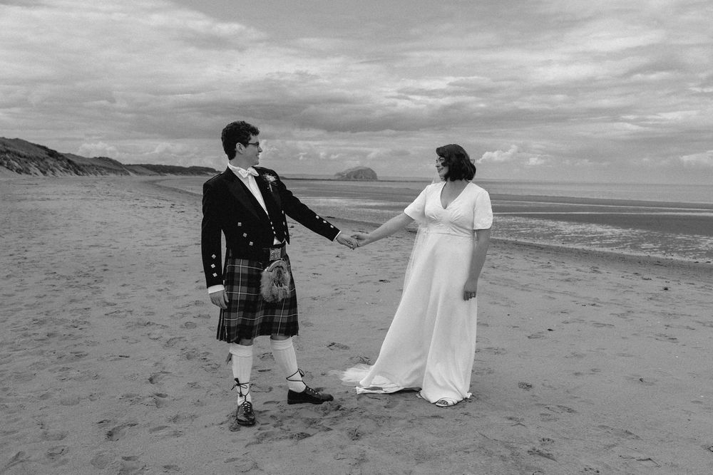 A couple in traditional Scottish and white wedding attire hold hands on a scenic beach during an overcast day.