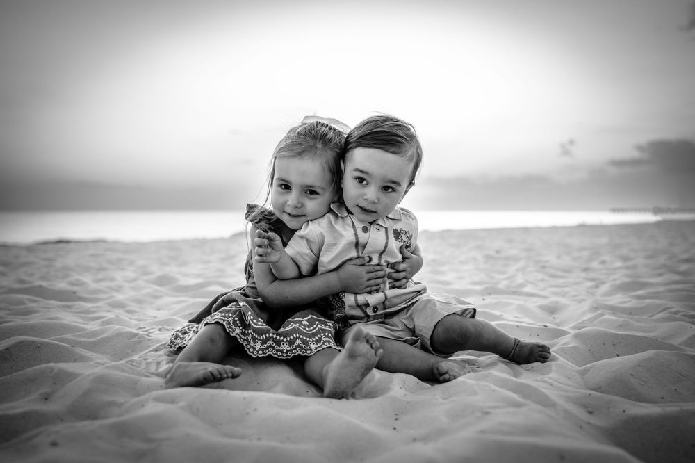 Two young children sit close together on a sandy beach in black and white photography.