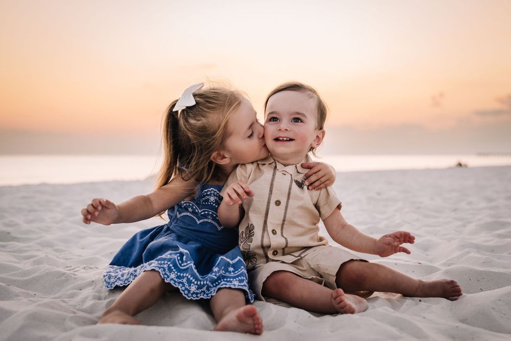 Two small children in casual beachwear sitting together on sandy beach at sunset with ocean in background.