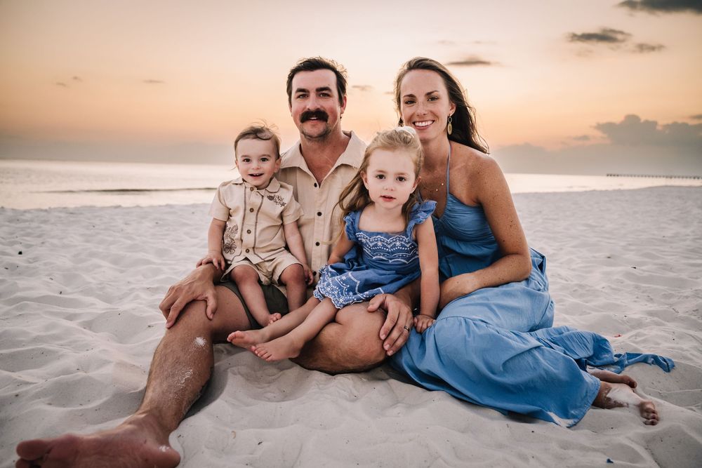 Family dressed in blue and beige outfits relaxing on beach sand during golden hour.