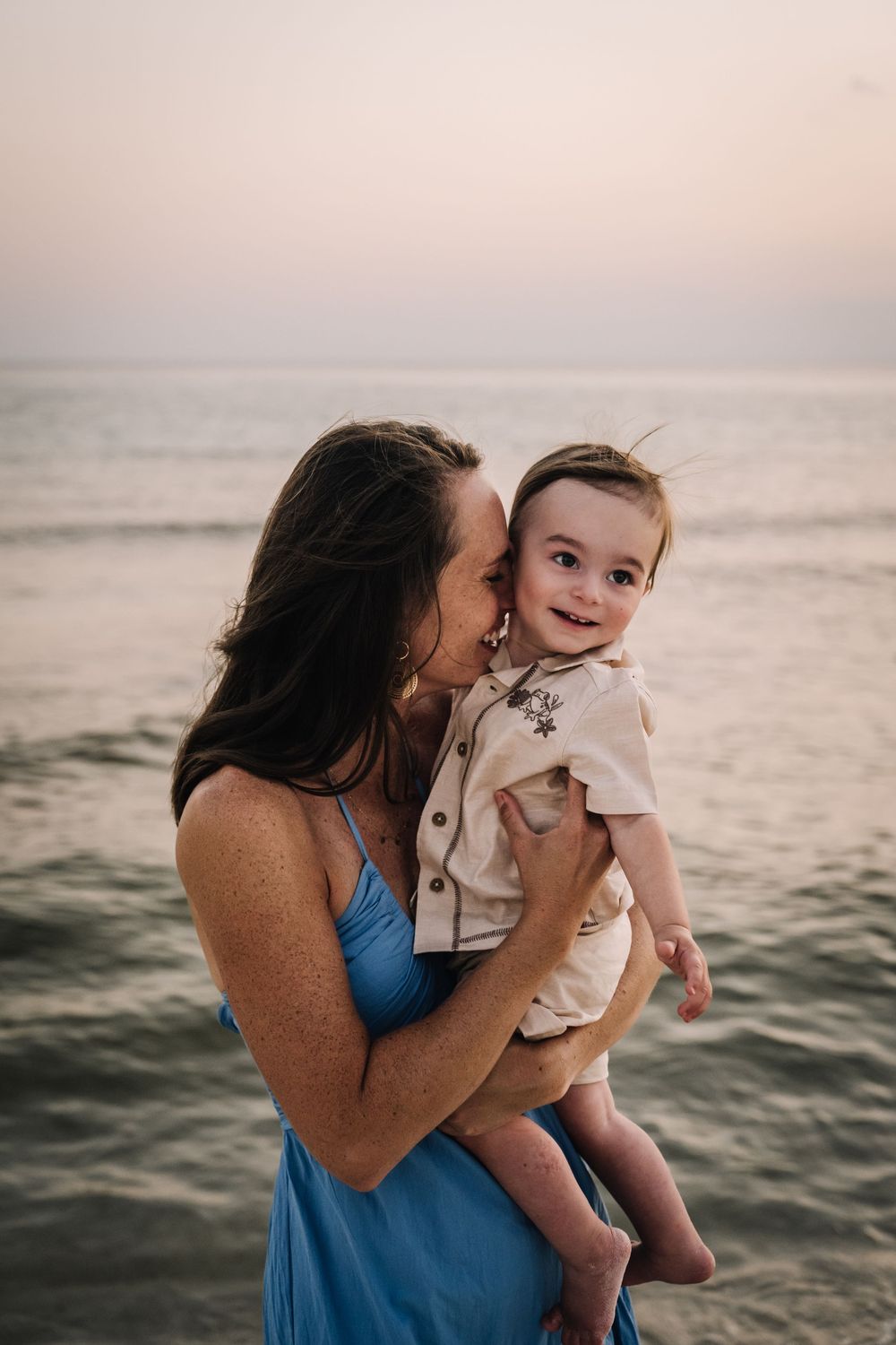 A tender moment captured at sunset by the ocean as a woman in a blue dress holds a smiling child.