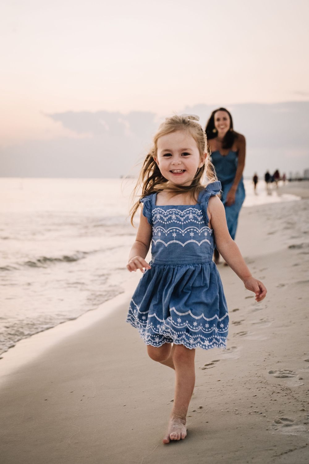 A young girl in a blue embroidered dress plays and runs along a sandy beach at sunset.