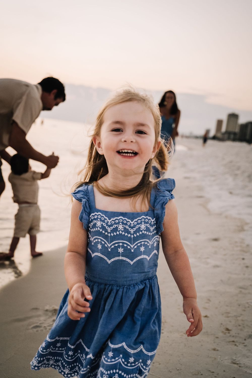 A child in a blue embroidered dress plays joyfully on a sandy beach at sunset.