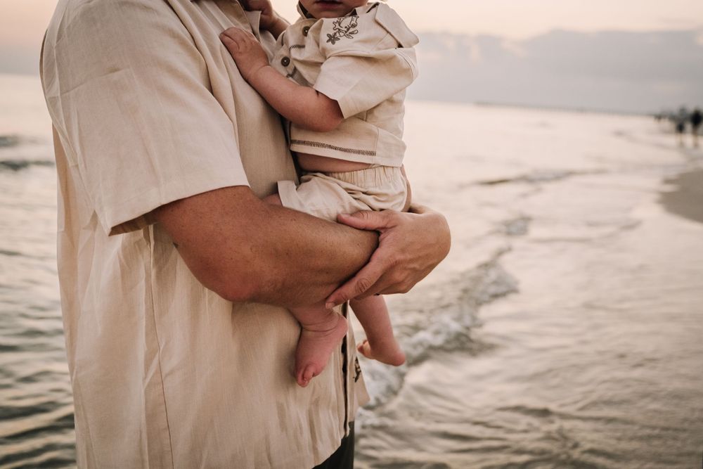 A couple holds their baby while standing on a beach at sunset in matching neutral colored outfits.