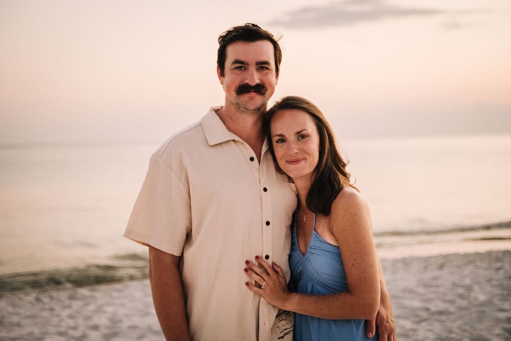 A couple embraces on the beach during sunset wearing casual attire in warm lighting.