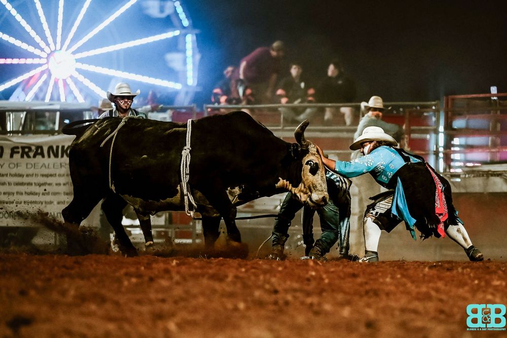 Bull wrestling event at night rodeo with illuminated carnival rides in background.