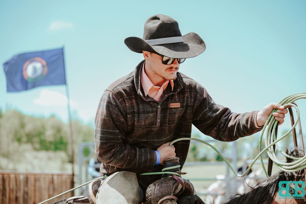 A cowboy in sunglasses and hat rides a horse in front of a blue sky with flags waving.