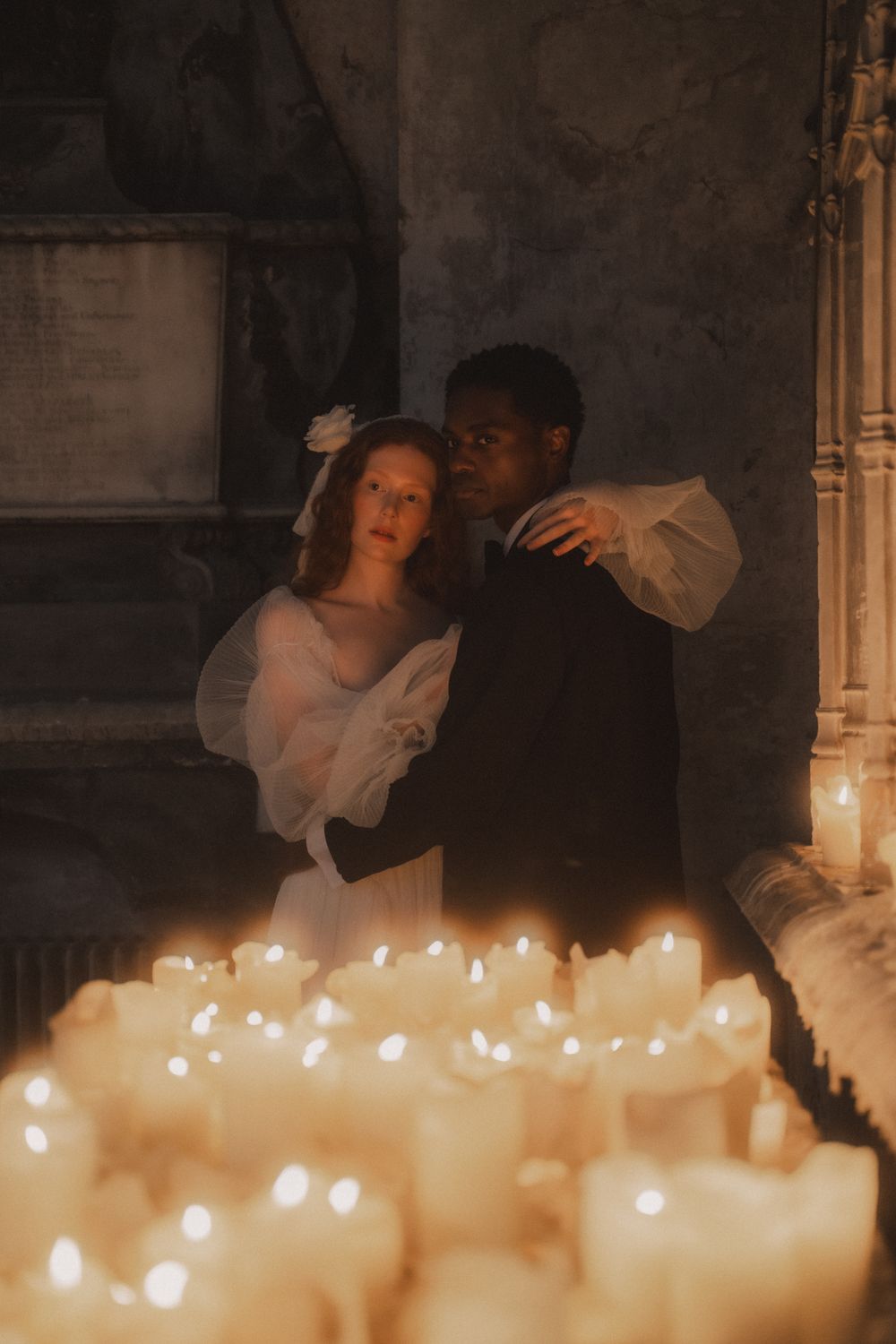 A romantic scene with glowing candles in the foreground while a couple embraces in soft shadows behind them.