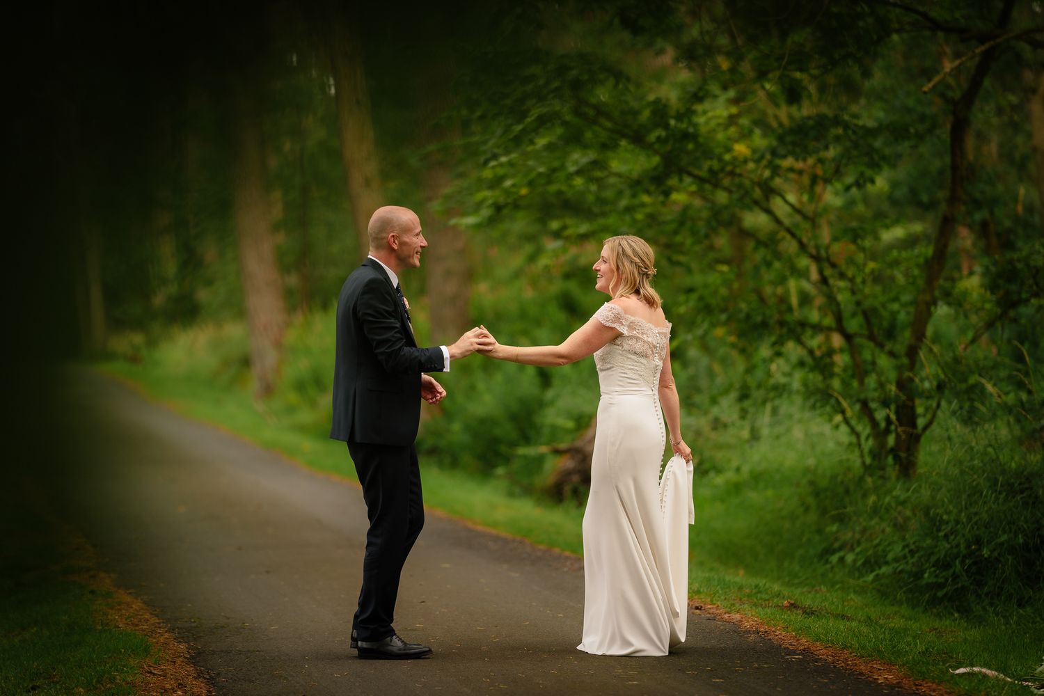 A couple in wedding attire poses together on a scenic forest pathway. Woodhill Hall.