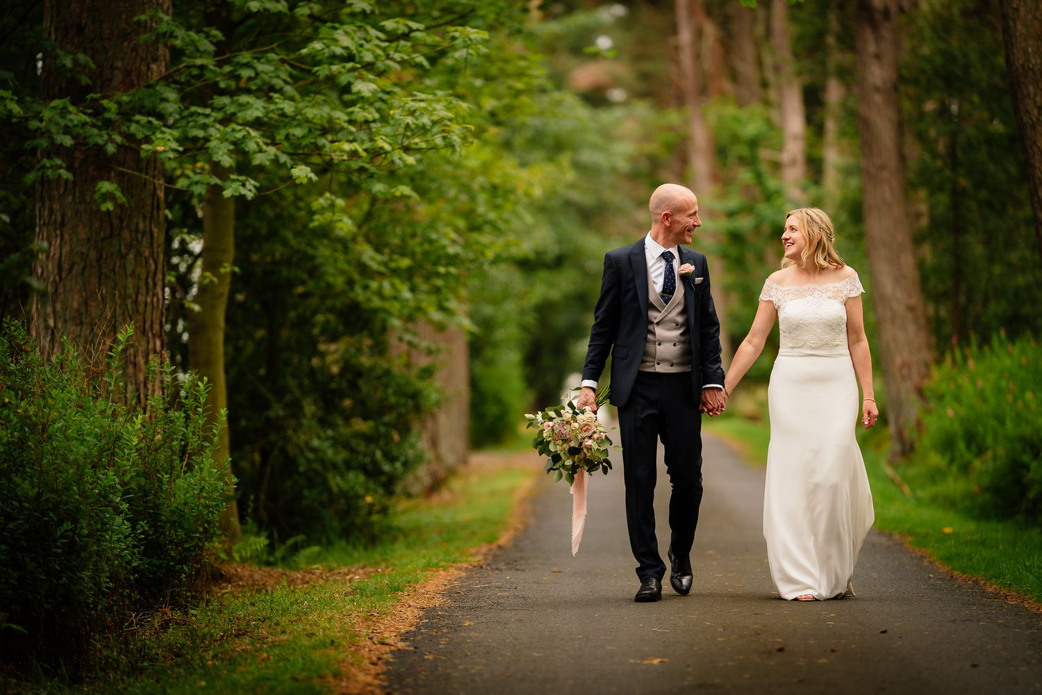 Tree-lined pathway with wedding couple walking in the distance surrounded by lush greenery. Woodhill Hall.