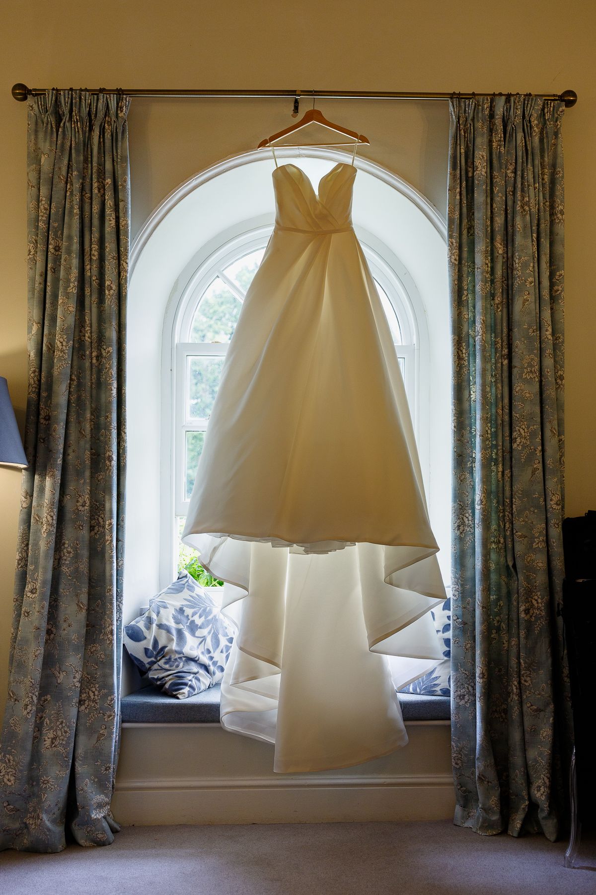 A white wedding dress hangs elegantly in front of an arched window with floral curtains, creating a romantic bridal scene.